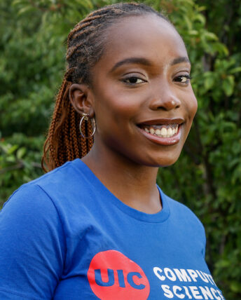 Tomi Okunola poses in a royal blue UIC computer science t-shirt against a background of green leaves